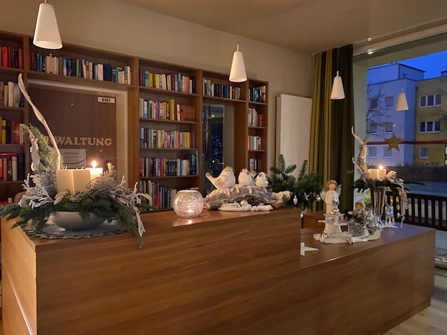 Stimmungsvoll beleuchteter Empfangsbereich mit Holztheke, weihnachtlicher Dekoration und Kerzenlicht. Im Hintergrund ein Bücherregal mit der Aufschrift „VERWALTUNG“ sowie Fenster mit Blick nach draußen – die Szene wirkt gemütlich und festlich.